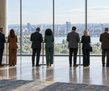Synthetic editorial illustration. A conference foyer. Attendees seen from behind stand in pairs at tall coffee tables facing broad windows, with plain blank banners at either side.