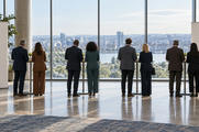Synthetic editorial illustration. A conference foyer. Attendees seen from behind stand in pairs at tall coffee tables facing broad windows, with plain blank banners at either side.