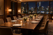 Synthetic editorial illustration. A private dining room at night. A long table is set with plain plates, glasses, folded napkins and small floral arrangements, with warm lamps and distant city lights beyond the windows.