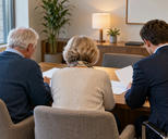 Synthetic editorial illustration. An advisory office. A rear-facing older couple and a suited adviser sit side by side at a wooden desk examining blank sheets, with plants, a lamp and a plain wall picture behind.