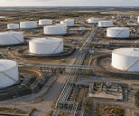 Synthetic editorial illustration. An oil storage facility viewed from above. White cylindrical tanks sit within containment berms beside a central corridor of pipelines and service roads, with flat countryside beyond.