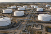 Synthetic editorial illustration. An oil storage facility viewed from above. White cylindrical tanks sit within containment berms beside a central corridor of pipelines and service roads, with flat countryside beyond.
