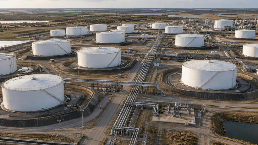Synthetic editorial illustration. An oil storage facility viewed from above. White cylindrical tanks sit within containment berms beside a central corridor of pipelines and service roads, with flat countryside beyond.