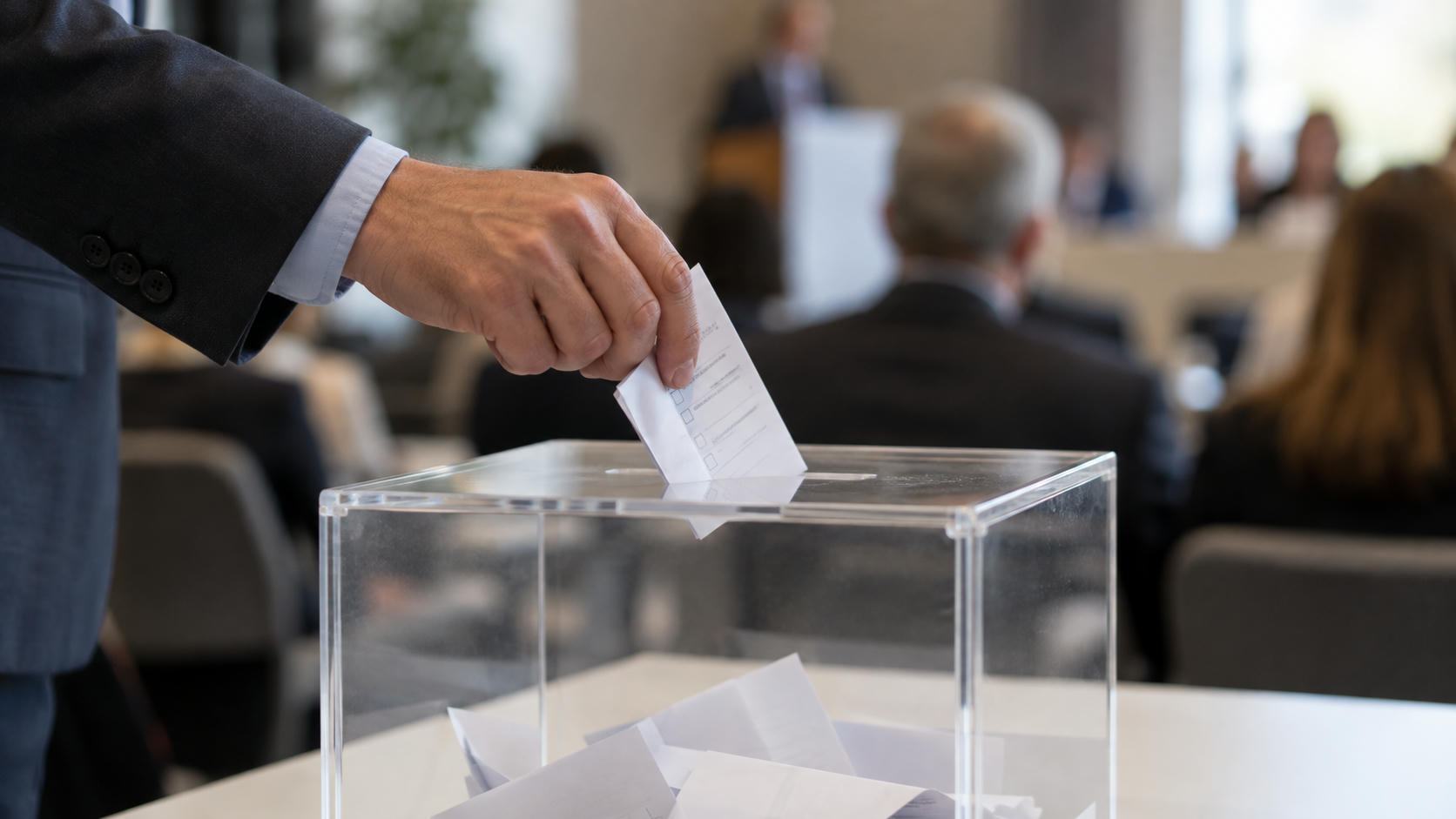 Synthetic editorial illustration. An adult hand places a partly folded populated ballot into a transparent box containing several papers.