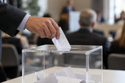 Synthetic editorial illustration. An adult hand places a partly folded populated ballot into a transparent box containing several papers.