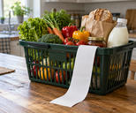 Synthetic editorial illustration. A green shopping basket filled with vegetables, bread, a plain jar and an unlabeled milk bottle sits on a wooden kitchen table. A long completely blank receipt hangs over the rim in window light.