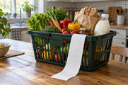 Synthetic editorial illustration. A green shopping basket filled with vegetables, bread, a plain jar and an unlabeled milk bottle sits on a wooden kitchen table. A long completely blank receipt hangs over the rim in window light.