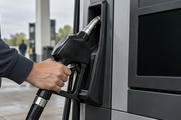 Synthetic editorial illustration. A hand grips a plain black fuel nozzle beside a grey pump. The large rectangular display is completely dark and blank, with the forecourt softly blurred beyond.