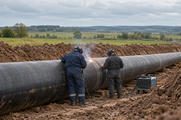 Synthetic editorial illustration. A countryside pipeline trench. Two workers in protective coveralls and welding helmets seen from behind weld a joint on a large steel pipe, with fields and a low treeline beyond.