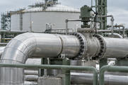 Synthetic editorial illustration. A large insulated metal pipe runs through a flanged control valve above fixed supports and a safety rail. A broad cylindrical storage tank and further process pipes stand behind it.
