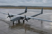Synthetic editorial illustration. An airbase apron. An unmarked grey twin-propeller drone has a high straight wing, two separated tail booms and upright fins joined by a horizontal tailplane, parked on wet concrete beneath an overcast sky.