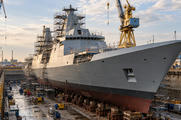 Synthetic editorial illustration. A shipyard dry dock. A large grey naval hull rests on supports surrounded by scaffolding, equipment and wet concrete, with tall cranes rising above its unfinished superstructure.