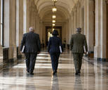 Synthetic editorial illustration. An institutional corridor. Three rear-facing officials carry thick closed folders beneath an arched ceiling, with tall windows, stone columns and pendant lamps reflected on the polished floor.