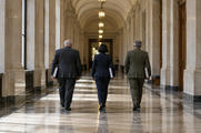 Synthetic editorial illustration. An institutional corridor. Three rear-facing officials carry thick closed folders beneath an arched ceiling, with tall windows, stone columns and pendant lamps reflected on the polished floor.