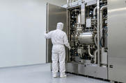 Synthetic editorial illustration. A semiconductor cleanroom. A worker in white protective coveralls faces an open metal service door. Polished cylindrical chambers, pipes and hoses fill the machine beside a plain pale wall.