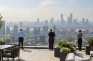 Synthetic editorial illustration. Three rear-facing workers stand at a rooftop glass railing above a hazy city skyline. Planters and outdoor seating occupy the sunlit terrace.