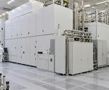 Synthetic editorial illustration. A semiconductor cleanroom. Huge white enclosed machines and polished pipework fill the hall, with two technicians in hooded white suits seen from behind beside dark monitors.