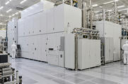 Synthetic editorial illustration. A semiconductor cleanroom. Huge white enclosed machines and polished pipework fill the hall, with two technicians in hooded white suits seen from behind beside dark monitors.