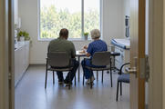Synthetic editorial illustration. A clinic consultation room. A patient and nurse seen from behind sit side by side as the nurse demonstrates a plain injection pen over a training pad on a small table.