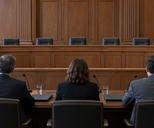 Synthetic editorial illustration. Three seated people viewed from behind face an empty raised bench in a wood-panelled hearing room. Microphones, closed folders and water glasses sit on the polished table in daylight.
