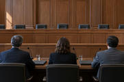 Synthetic editorial illustration. Three seated people viewed from behind face an empty raised bench in a wood-panelled hearing room. Microphones, closed folders and water glasses sit on the polished table in daylight.