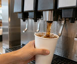 Synthetic editorial illustration. A restaurant drink station. A cropped hand holds a blank white paper cup under a stainless-steel fountain nozzle as a brown fizzy drink pours over ice; plain dispenser panels and a drip tray fill the close view.