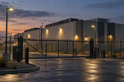 Synthetic editorial illustration. A secured data-centre facility stands behind a tall metal fence and closed sliding gate. Rooftop cooling equipment and warm exterior lamps are visible under an evening sky.