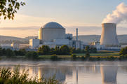 Synthetic editorial illustration. A generic domed power-station building and cooling tower are framed across a river at dawn.