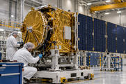 Synthetic editorial illustration. Two cleanroom workers work beside a supported gold-toned satellite assembly with a large blue panel in a high bay.