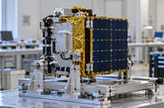 Synthetic editorial illustration. A compact satellite-like hardware assembly with a blue panel and gold-toned blanket is supported on an integration fixture.