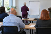 Synthetic editorial illustration. Varied office staff viewed from behind sit facing a white-haired trainer in a plum suit beside a blank flipchart. Plain closed laptops and blank notebooks rest on tables in a bright training room.