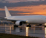 Synthetic editorial illustration. An airport runway. A plain white cargo jet taxis across wet pavement under an orange dusk sky, with landing lights and blue airport lights reflecting around its wheels.