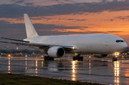 Synthetic editorial illustration. An airport runway. A plain white cargo jet taxis across wet pavement under an orange dusk sky, with landing lights and blue airport lights reflecting around its wheels.