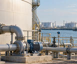 Synthetic editorial illustration. A mounted pump and flange-connected pipework sit beside a storage tank at a waterfront terminal.