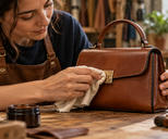 Synthetic editorial illustration. An artisan steadies a small plain leather handbag while polishing its metal clasp with a soft cloth.