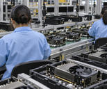 Synthetic editorial illustration. Two workers in blue anti-static coats viewed from behind assemble plain graphics cards along a conveyor. Black fans, metal heat sinks and component carriers fill the foreground, with dark monitors beyond.
