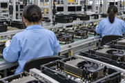 Synthetic editorial illustration. Two workers in blue anti-static coats viewed from behind assemble plain graphics cards along a conveyor. Black fans, metal heat sinks and component carriers fill the foreground, with dark monitors beyond.