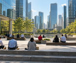 Synthetic editorial illustration. Rear-facing workers eat from small containers on steps and benches in a sunlit plaza. Trees, a reflecting pool and glass towers surround the open space.