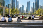 Synthetic editorial illustration. Rear-facing workers eat from small containers on steps and benches in a sunlit plaza. Trees, a reflecting pool and glass towers surround the open space.