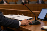 Synthetic editorial illustration. A cropped adult presses a hearing-desk microphone button beside an open evidence binder and powered tablet.