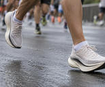 Synthetic editorial illustration. A runner's plain light-coloured mesh shoes with thick curved soles are caught mid-stride on a wet city road. Other runners' lower legs blur behind in a low ground-level view.