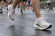 Synthetic editorial illustration. A runner's plain light-coloured mesh shoes with thick curved soles are caught mid-stride on a wet city road. Other runners' lower legs blur behind in a low ground-level view.