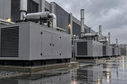 Synthetic editorial illustration. A data-centre service yard contains a row of grey backup-generator enclosures. Tall exhaust stacks rise beside the building as rainwater reflects equipment and lamps.