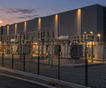 Synthetic editorial illustration. A substation stands beside a windowless data-centre building. Large transformers and switchgear occupy a fenced gravel yard under warm lamps at dusk.