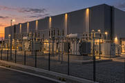 Synthetic editorial illustration. A substation stands beside a windowless data-centre building. Large transformers and switchgear occupy a fenced gravel yard under warm lamps at dusk.