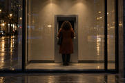 Synthetic editorial illustration. An ATM lobby at night. A person in a rust coat seen from behind stands at a single cash machine with a dark screen, while wet street lights reflect across the surrounding glass.