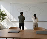 Synthetic editorial illustration. Two people seen from behind face a whiteboard of circles and arrows in a sunlit office. Two closed laptops rest on the wooden table beside a potted tree.