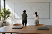 Synthetic editorial illustration. Two people seen from behind face a whiteboard of circles and arrows in a sunlit office. Two closed laptops rest on the wooden table beside a potted tree.