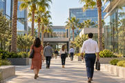 Synthetic editorial illustration. Employees of varied builds viewed from behind walk along a sunny path between glass office buildings. Palm trees and planted beds line the quiet campus.