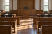 Synthetic editorial illustration. An empty wood-panelled courtroom contains rows of benches, plain tables and a raised judge's seat. A small gavel rests on the bench while daylight from tall windows falls across the floor.